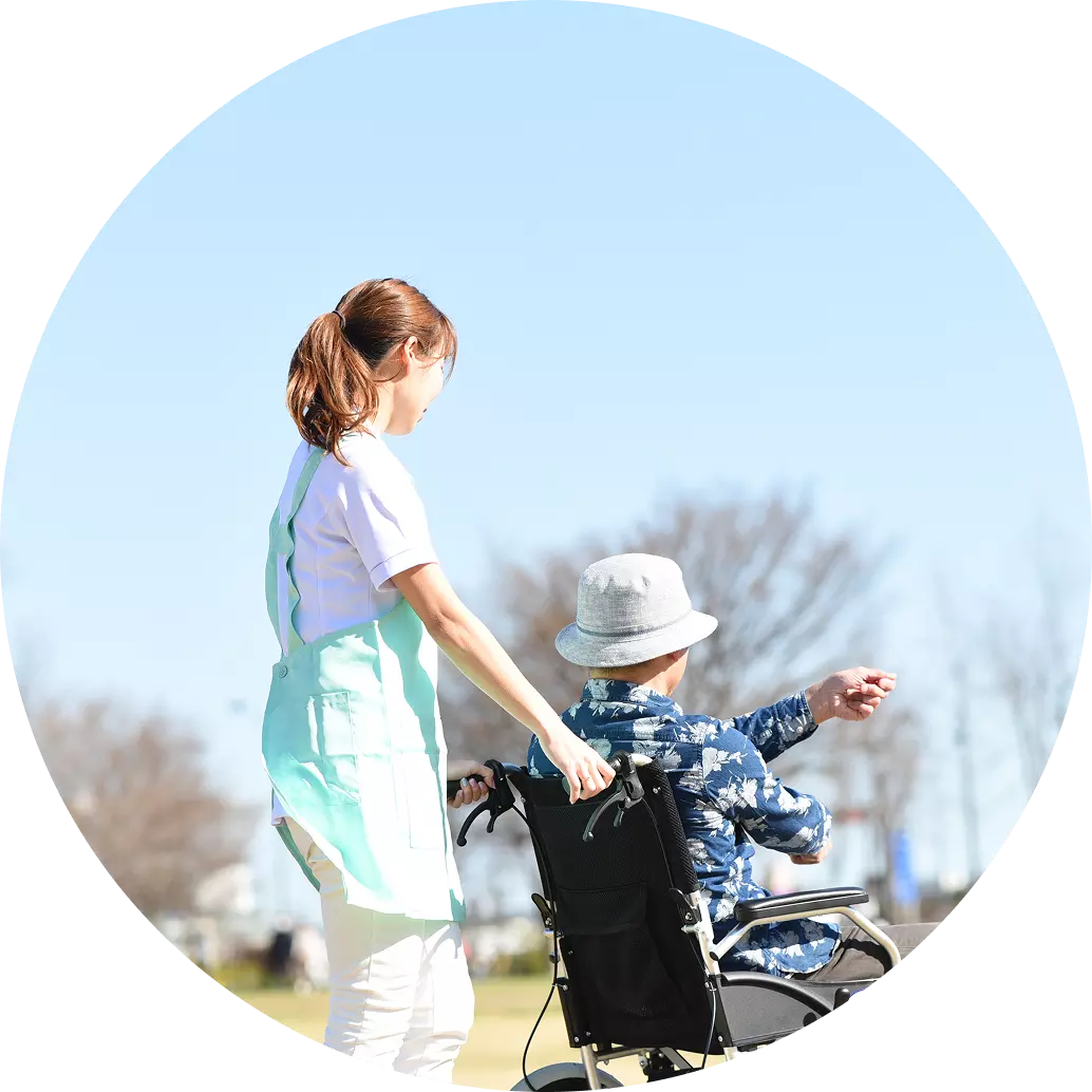 屋外の広場で介護士の人が車いすに乗った人と一緒に車いすを押しながら歩いている画像